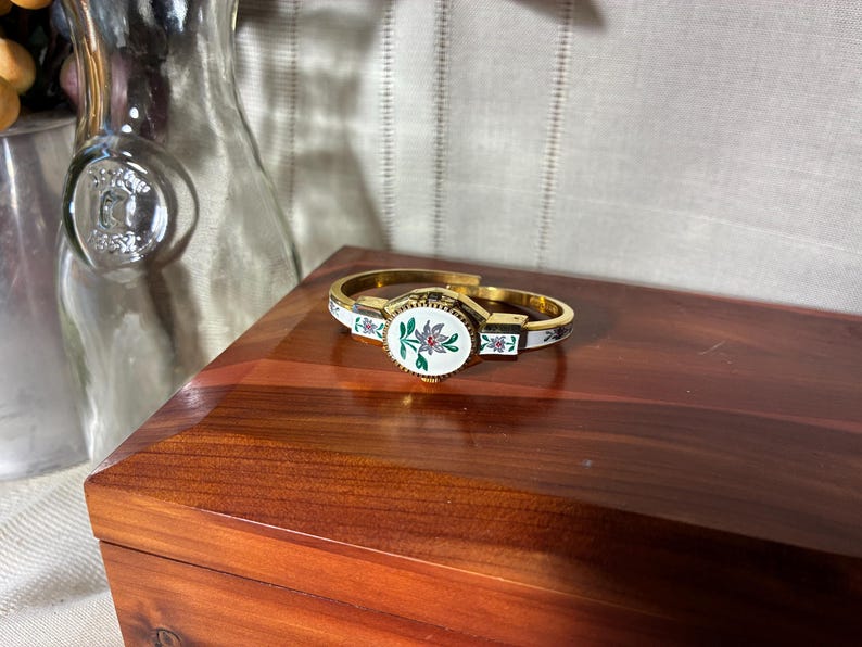 May include: Gold-toned hinged bracelet with a central oval design featuring a floral motif in pink and green on a white background. The bracelet has white enamel accents with floral details. A vintage jewelry piece.