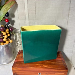 May include: A square, emerald green ceramic vase with a yellow interior. The vase is displayed on a wooden surface, with a metal pitcher of faux berries and a glass jar in the background. The vase is a decorative item.