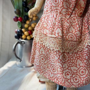 May include: A doll wearing a dress made of light-colored fabric with a repeating orange floral pattern. The dress features a tiered skirt design with a decorative beige lace trim along the horizontal seam of the middle ruffle layer.