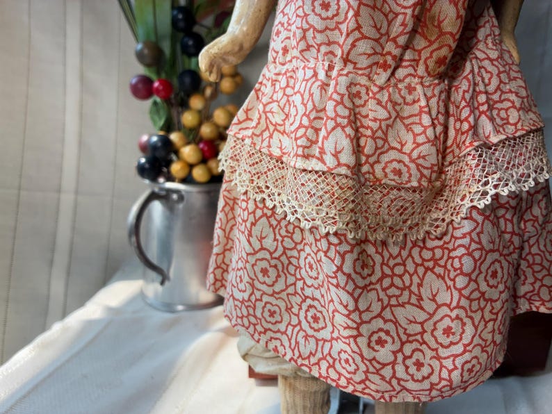May include: A doll wearing a dress made of light-colored fabric with a repeating orange floral pattern. The dress features a tiered skirt design with a decorative beige lace trim along the horizontal seam of the middle ruffle layer.