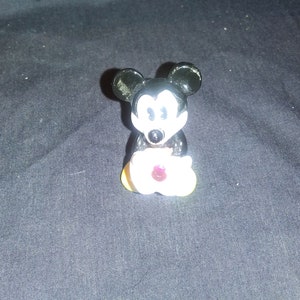May include: A small, black and white Mickey Mouse figurine holding a white flower with a red center. The figurine has large black ears and a smiling face. The flower has yellow petals.