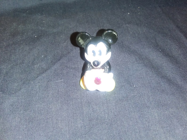 May include: A small, black and white Mickey Mouse figurine holding a white flower with a red center. The figurine has large black ears and a smiling face. The flower has yellow petals.