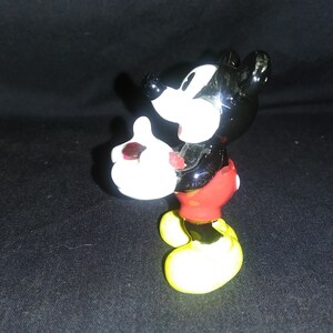 May include: A ceramic Mickey Mouse figurine. The figure has a black body, white gloves, red shorts, and yellow shoes. The figurine is posed with one hand raised and the other holding a small object.