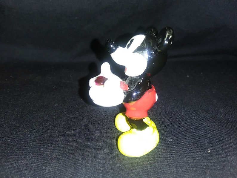 May include: A ceramic Mickey Mouse figurine. The figure has a black body, white gloves, red shorts, and yellow shoes. The figurine is posed with one hand raised and the other holding a small object.