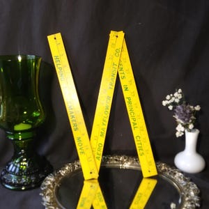 May include: A yellow folding ruler with black markings and text, including "Helping Makers Move" and "Cents in Principal Cities." The ruler is positioned on a silver tray, with a green glass and a small white vase with flowers in the background.
