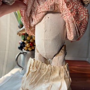May include: A vintage doll with a pale, molded composition torso and limbs. The doll wears a gathered, cream-colored fabric pantaloons and a skirt featuring a red floral pattern on a light background. The construction shows visible seams.