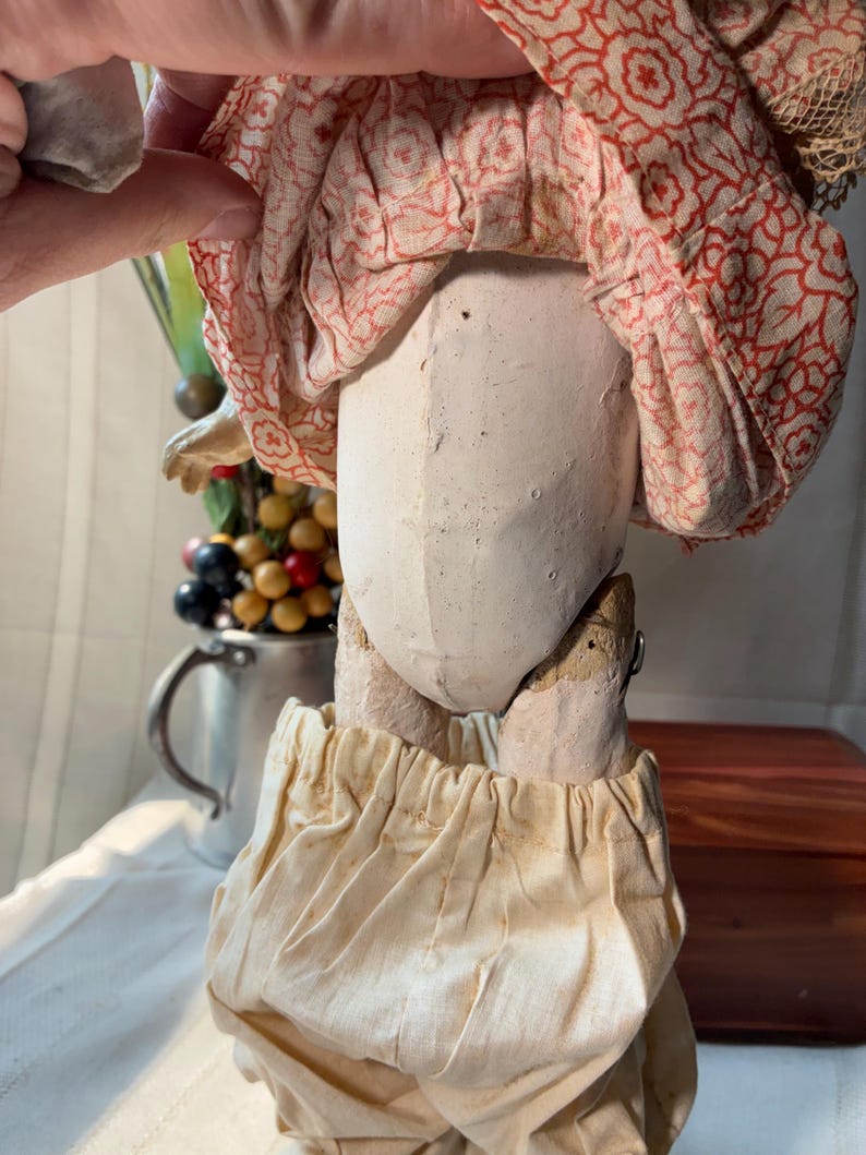 May include: A vintage doll with a pale, molded composition torso and limbs. The doll wears a gathered, cream-colored fabric pantaloons and a skirt featuring a red floral pattern on a light background. The construction shows visible seams.