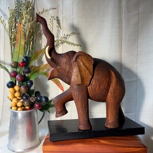 May include: A brown wooden elephant sculpture with a raised trunk and one front leg lifted, featuring a single white tusk. The elephant is mounted on a rectangular black base, showcasing detailed carved textures across its body and ears.