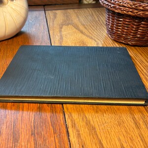 May include: A closed, rectangular black book with a textured cover. The edges of the pages are a golden yellow colour. The book is lying on a wooden surface.