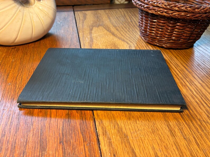 May include: A closed, rectangular black book with a textured cover. The edges of the pages are a golden yellow colour. The book is lying on a wooden surface.