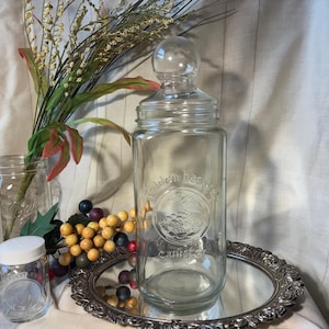 May include: A clear glass canister with a round lid and a decorative emblem, sitting on a silver-colored tray. The canister has the words "Golden Harvest" and "canister" etched on it. Other glass jars and decorative elements are in the background.