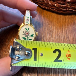 May include: A vintage gold-toned watch with a white face featuring a floral design with red and green accents. The watch is approximately 1.5 inches in length, as measured by a yellow tape measure.