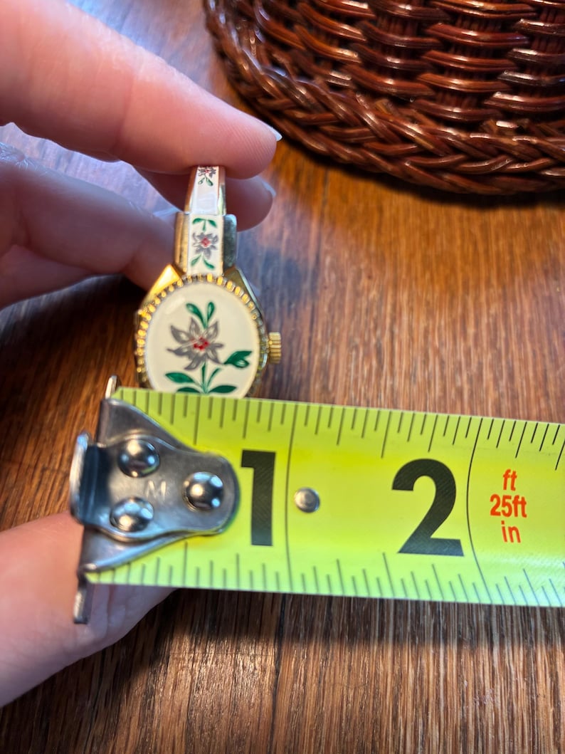 May include: A vintage gold-toned watch with a white face featuring a floral design with red and green accents. The watch is approximately 1.5 inches in length, as measured by a yellow tape measure.