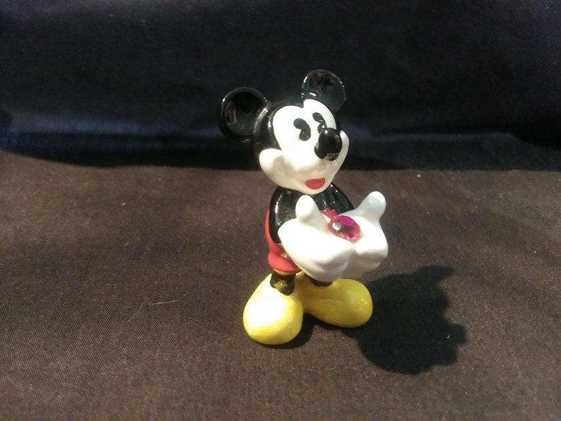 May include: A small, ceramic Mickey Mouse figurine. Mickey is depicted in his classic attire: black ears and body, white gloves, red shorts, and yellow shoes. He holds a pink gem in his hands.