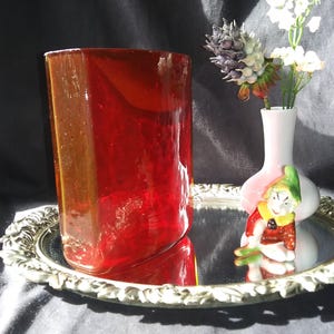 May include: A red glass vase with a textured surface sits on a silver tray. A small white vase with a pink and green clown design holds a bouquet of white flowers.