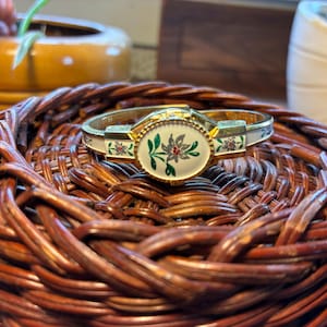 May include: A gold-toned hinged bracelet with an oval floral design in the center. The bracelet has white enamel accents with red and green floral details. It rests on a woven brown basket.