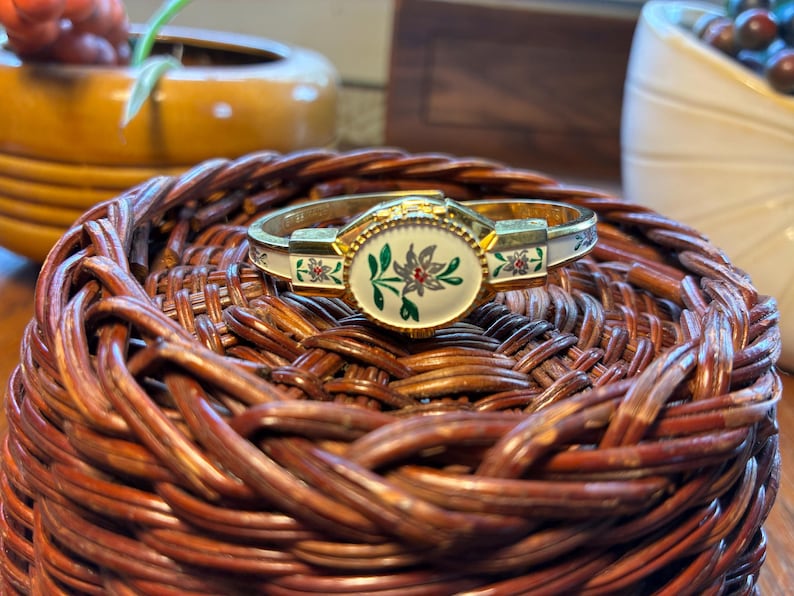 May include: A gold-toned hinged bracelet with an oval floral design in the center. The bracelet has white enamel accents with red and green floral details. It rests on a woven brown basket.