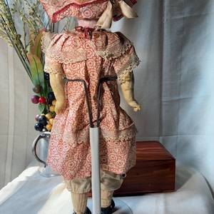May include: A vintage doll viewed from the back, wearing a dress with a red floral pattern on a cream background, lace trim, and a matching bonnet. The doll is supported by a metal stand and wears beige trousers and black shoes.