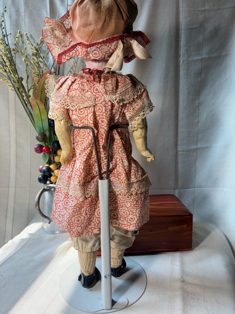 May include: A vintage doll viewed from the back, wearing a dress with a red floral pattern on a cream background, lace trim, and a matching bonnet. The doll is supported by a metal stand and wears beige trousers and black shoes.