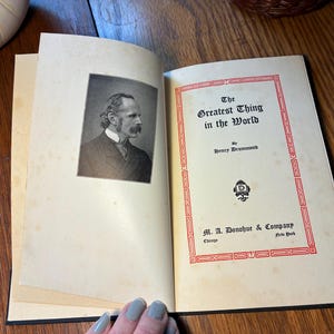 May include: An open book titled "The Greatest Thing in the World" by Henry Drummond. The cover has a red border and the publisher's details. The left page displays a black and white portrait of a man.