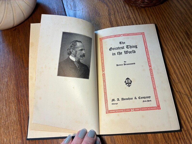 May include: An open book titled "The Greatest Thing in the World" by Henry Drummond. The cover has a red border and the publisher's details. The left page displays a black and white portrait of a man.