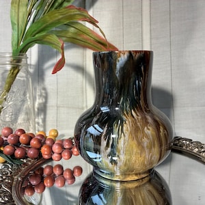 May include: A decorative ceramic vase with a glossy finish, featuring a blend of brown, black, and gold tones. The vase has a rounded body and a flared opening. It sits on a silver-colored tray, with a glass jar and faux berries in the background.