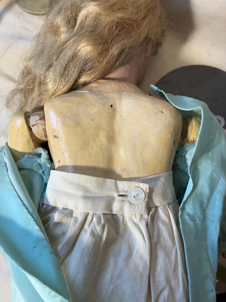 Op de afbeelding: Een vintage pop met blond haar en een lichte huidskleur. De pop draagt een witte rok en een lichtblauwe top. De achterkant van de pop is zichtbaar, met de houten gewrichten en de knoopsluiting op de rok.