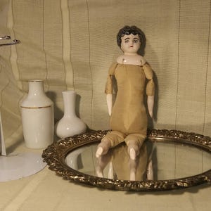 May include: A porcelain doll with black hair and a brown dress sits on a gold-colored tray with a mirror. The doll has a painted face with blue eyes and red lips.