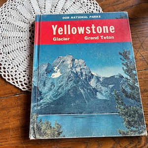 May include: Vintage book titled "Yellowstone Glacier Grand Teton" from the "Our National Parks" series. The cover features a scenic mountain and lake landscape with a red and blue color scheme. The book is placed on a wooden surface next to a white lace doily.