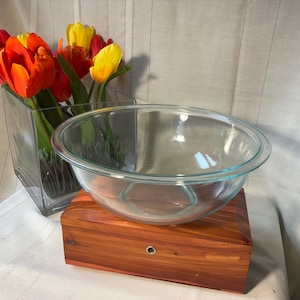 May include: A clear glass mixing bowl with a wide rim sits atop a rectangular wooden box with a visible metal grommet. Next to the bowl is a square glass vase containing a bouquet of vibrant red, orange, and yellow tulip flowers with green stems.
