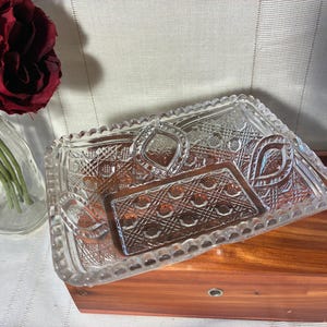 May include: A rectangular clear pressed glass dish featuring a decorative scalloped edge and intricate geometric patterns including cross-hatching and diamond shapes. The glass tray has a central panel with a grid design and two oval motifs.