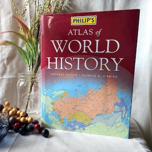 May include: A hardcover book titled "PHILIP'S ATLAS of WORLD HISTORY" with a red cover and a world map illustration. The book is displayed with decorative elements including a glass jar, a small glass bottle, and faux grapes.