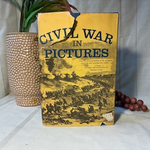 May include: A vintage book titled "Civil War in Pictures" with a yellow cover and black text. The cover features a battle scene illustration. The book is slightly damaged, with a torn top edge. A decorative vase and faux grapes are in the background.