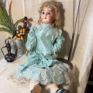 Op de afbeelding: Een vintage porseleinen pop met blond haar, gekleed in een lichtblauwe satijnen jurk met kanten rand. De pop heeft roze wangen en zit. De pop draagt witte sokken en zwarte schoenen.