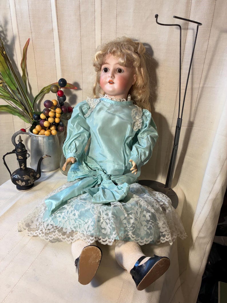 Op de afbeelding: Een vintage porseleinen pop met blond haar, gekleed in een lichtblauwe satijnen jurk met kanten rand. De pop heeft roze wangen en zit. De pop draagt witte sokken en zwarte schoenen.