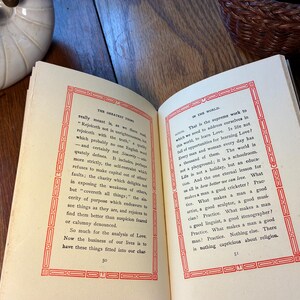May include: An open book with text on cream pages, framed by red borders. The book rests on a wooden surface. A white ceramic bowl and a woven basket are visible in the background.