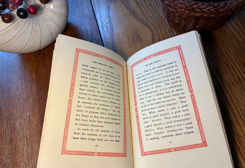 May include: An open book with text on cream pages, framed by red borders. The book rests on a wooden surface. A white ceramic bowl and a woven basket are visible in the background.