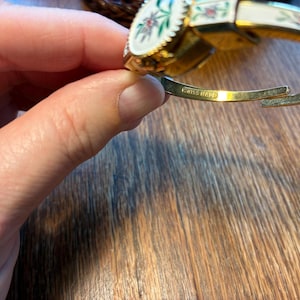 May include: A gold-toned hinged bracelet with a white enamel finish. The bracelet features floral designs in red, green, and white. The words "SWISS MADE" are engraved on the inside of the bracelet.