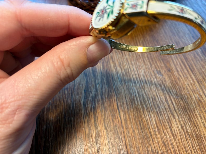 May include: A gold-toned hinged bracelet with a white enamel finish. The bracelet features floral designs in red, green, and white. The words "SWISS MADE" are engraved on the inside of the bracelet.