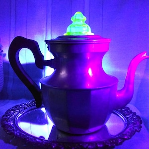 May include: A vintage purple teapot with a black handle and a green glass lid. The teapot sits on a silver-colored tray. The image is illuminated with purple and blue light.