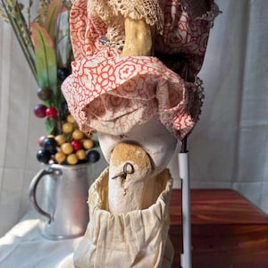 May include: A doll with a pale, textured head and torso, wearing a red and white floral patterned fabric garment with lace trim. The doll features a visible metal pin in the neck area and is dressed in a cream-colored fabric lower garment.