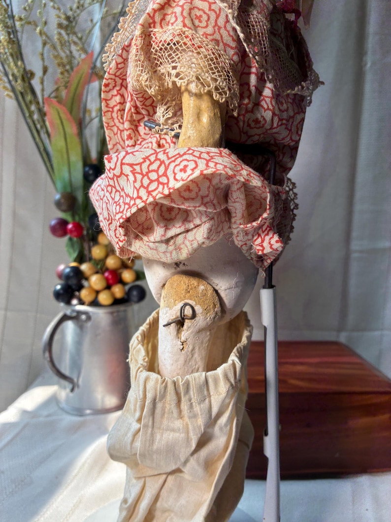 May include: A doll with a pale, textured head and torso, wearing a red and white floral patterned fabric garment with lace trim. The doll features a visible metal pin in the neck area and is dressed in a cream-colored fabric lower garment.