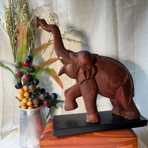 May include: A hand-carved wooden elephant figurine with a raised trunk, textured skin details, and visible tusks. The brown elephant stands on a rectangular black base, positioned next to a silver metal pitcher filled with decorative faux berries.