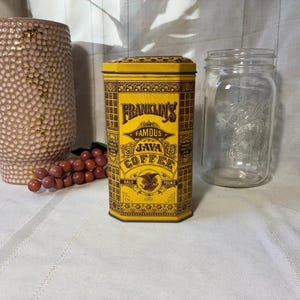 May include: A vintage yellow and brown Franklin's Famous Java Coffee tin. The tin has a rectangular shape with a decorative pattern and the words "Famous Java Coffee" in a stylized font. A clear glass jar and a decorative vase are in the background.
