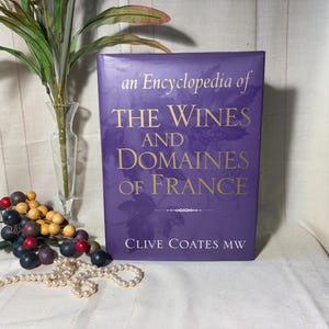 May include: A hardcover book titled "An Encyclopedia of The Wines and Domaines of France" in gold lettering on a purple background. The book is next to a clear glass vase with green leaves and a decorative cluster of berries.