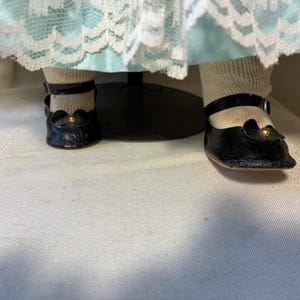 Op de afbeelding: Close-up van de voeten van een pop met zwarte leren Mary Jane-schoenen met goudkleurige accenten. De pop draagt ook witte sokken. De jurk van de pop is zichtbaar op de achtergrond, met een lichtblauwe rok en witte kanten afwerking.