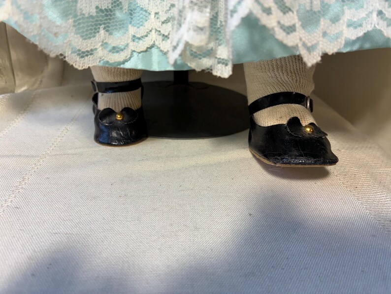 Op de afbeelding: Close-up van de voeten van een pop met zwarte leren Mary Jane-schoenen met goudkleurige accenten. De pop draagt ook witte sokken. De jurk van de pop is zichtbaar op de achtergrond, met een lichtblauwe rok en witte kanten afwerking.