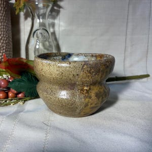 May include: A handmade ceramic bowl with a unique, textured surface. The bowl features a rounded base and a wider, slightly squared top. The color palette includes shades of tan, brown, and hints of blue, creating a rustic aesthetic. Ideal for serving or display.