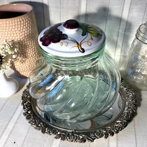 May include: A clear glass jar with a decorative lid featuring a grape design, set on a silver-colored tray. A pink textured vase and a Ball jar are also visible, suggesting a vintage or farmhouse theme.