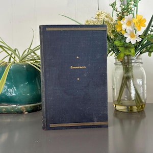 May include: A vintage book with a navy blue cover and gold accents. The title "Emerson" is printed in gold, with a star above and below the title. The book is standing upright on a gray surface.